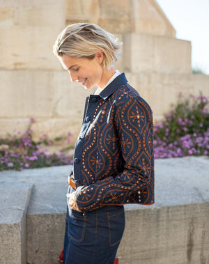 A woman stands indoors by large windows, wearing the MERSEA - FIFI JACKET—an indigo cotton jacket with cutwork embroidery—over a white shirt, paired with belted high-waisted blue pants and brown sandals. Sunlight creates grid-like shadows on the floor and wall.