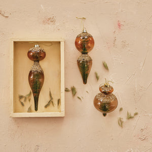 The CREATIVE COOP MICA FLAKES BOTTLE BRUSH TREE ORNAMENT set includes three vintage-style glass ornaments with gold hanging strings, brown-tinted glass, elongated shapes, and green pine trees inside.