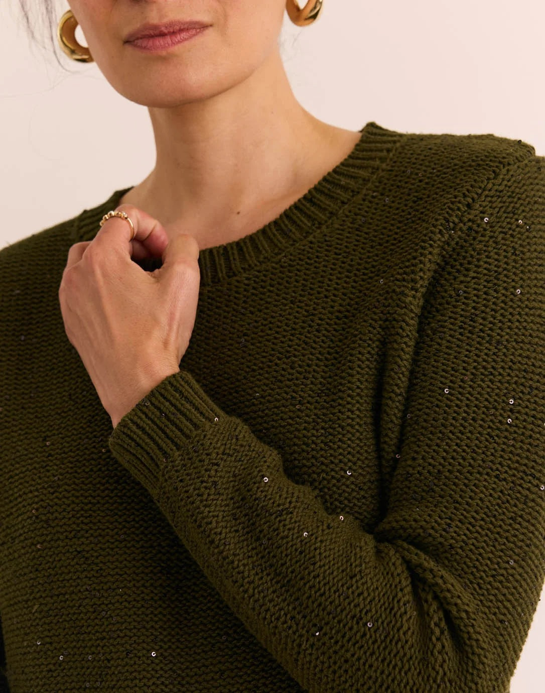 A person wearing the HINSON WU Queenie Long Sleeve Sequin Sweater in Oregano—an everyday essential crafted from breathable cotton with subtle sequin embellishments—paired with gold hoop earrings and touching their collar with one hand.