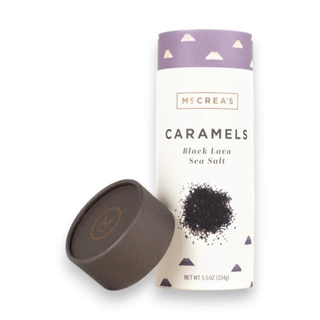With the lid removed, the MCCREA’S CANDIES - BLACK LAVA SEA SALT CARAMELS tube by McCREA’S CANDIES reveals its buttery caramel treats inside. The packaging is adorned with illustrations of Hawaiian black sea salt and bold purple mountain motifs.