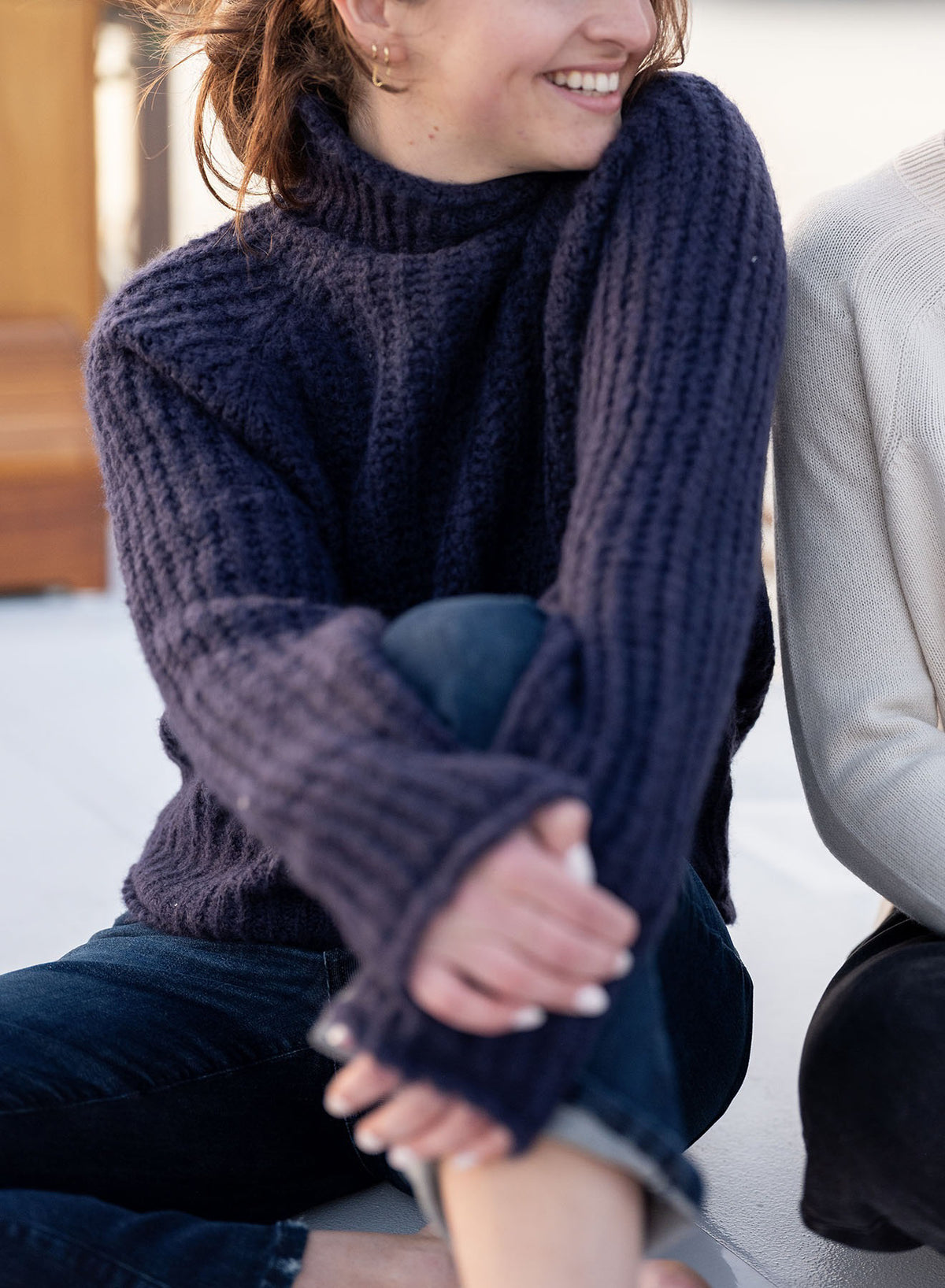 A person wearing the MERSEA - Beachside Softy Sweater in Navy and dark jeans sits with arms crossed over their knees, smiling and looking to the side. Next to them, only part of another person in a light sweater is visible.