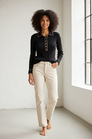 A person with curly hair stands barefoot indoors, smiling near a large window while wearing the MERSEA LUDLOW Long Sleeve Polo in Ink and light-colored jeans.