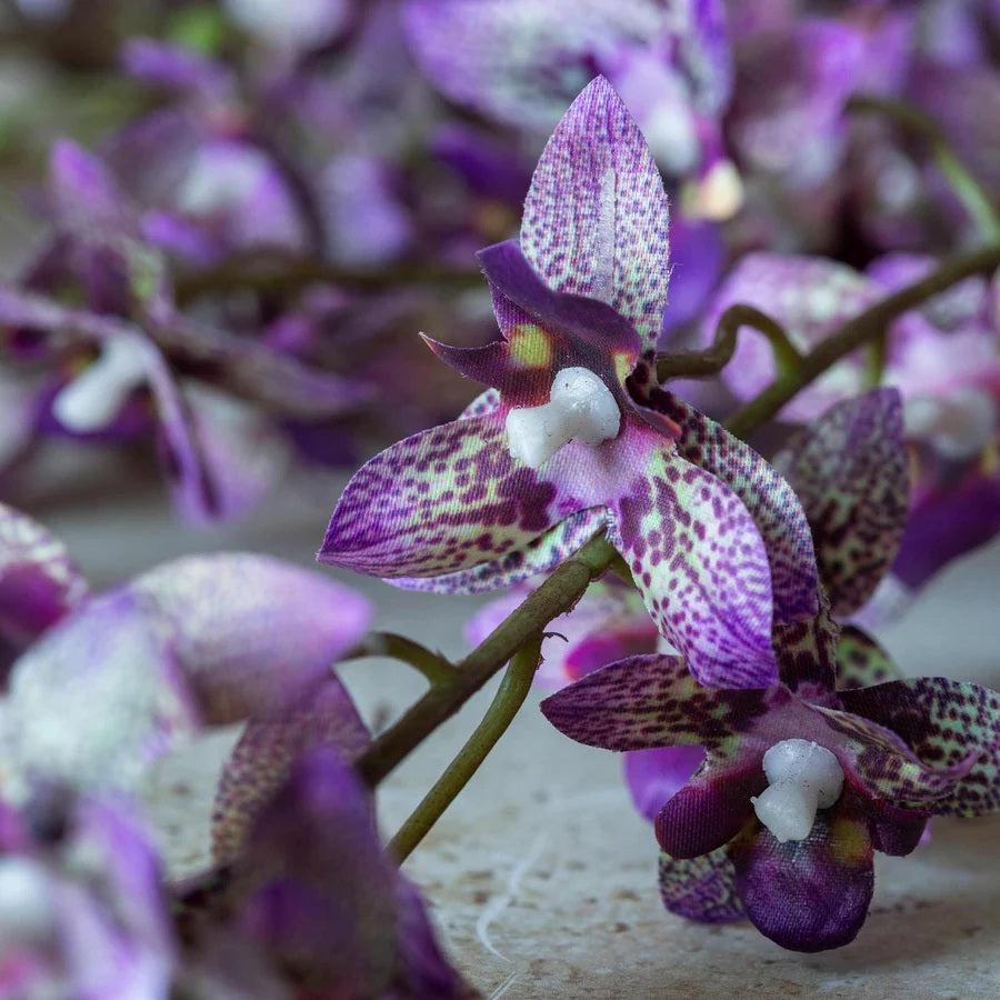 The ABIGAIL AHERN ONCIDIUM ORCHID is a single artificial stem featuring sparse small purple flowers and buds on delicate branches, presented against a plain white background.