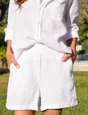Wearing FRANK & EILEEN's MAE LINEN SHORT, a person in a white button-up with rolled sleeves stands outdoors, hands in pockets; lush greenery and a pool behind evoke an effortless Amalfi Coast vibe.