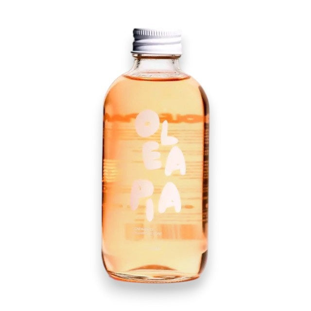 A clear glass bottle with a silver cap contains a light pink liquid. The label displays “OLEA PIA” in white, stylized letters. This elegant OLEA PIA - ROSE BALSAMIC VINEGAR, crafted in Italy, stands out against the plain white background.