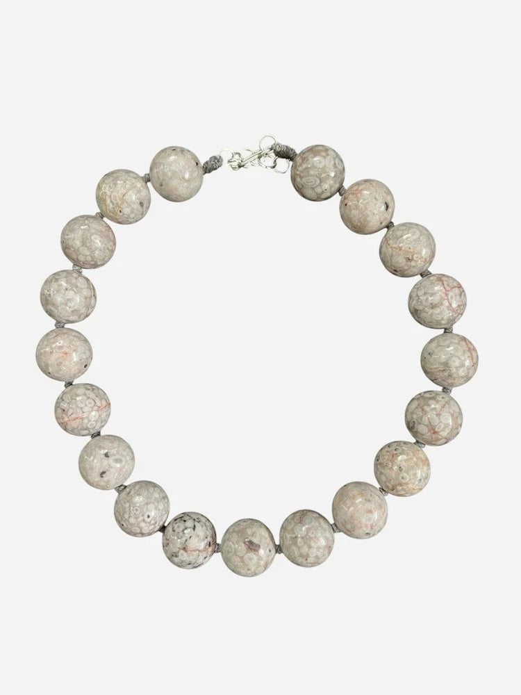 The ANN LIGHTFOOT Fossil Coral Ras du Cou Necklace features large, polished round fossil coral beads in light gray and beige with circular patterns and a sterling silver clasp, shown on a dark gray background.