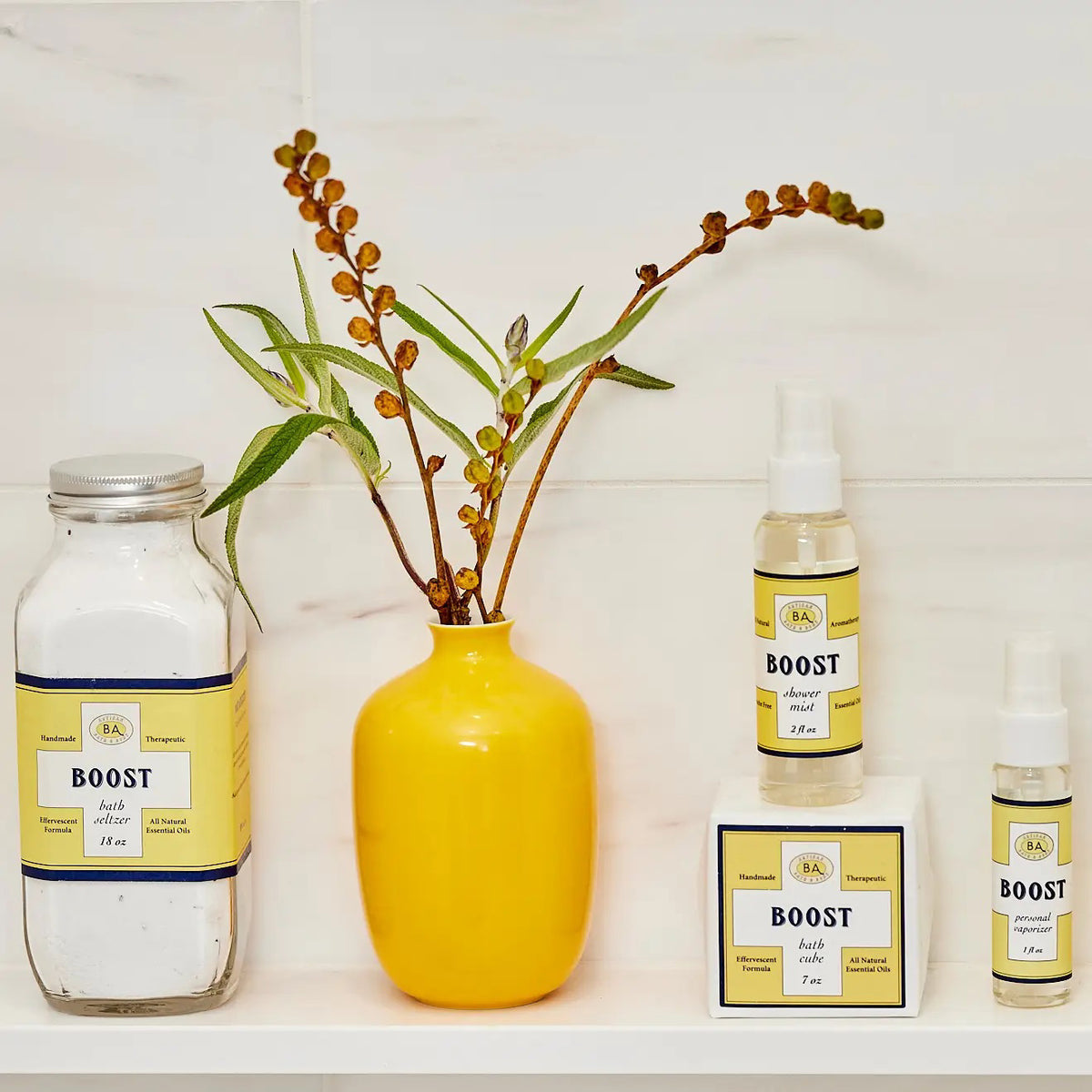 A collection of BAUDELAIRE&#39;s &quot;Boost&quot; skincare products, including bath salts, cleansing oil, and a peppermint-infused BAUDELAIRE - BOOST SHOWER MIST in a spray bottle, is displayed on a shelf alongside a tall yellow vase with green and brown stems.