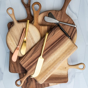 A stack of BE HOME - ACACIA RECTANGULAR BOARD by BE HOME, INC features three cheese knives—gold, copper, and silver—resting on top, set against a light geometric-patterned background.