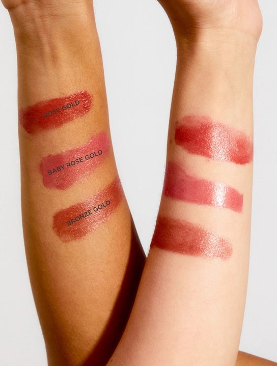 Two forearms display three shimmery lip swatches labeled Rose Gold, Baby Rose Gold, and Bronze Gold—each showing delicate tonal differences on various skin tones. The luminous rose gold shade from LANOLIPS 101 OINTMENT ROSE GOLD imparts a fresh, radiant look, similar to a hydrating cheek and lip stain or your favorite lanolin-based balm.