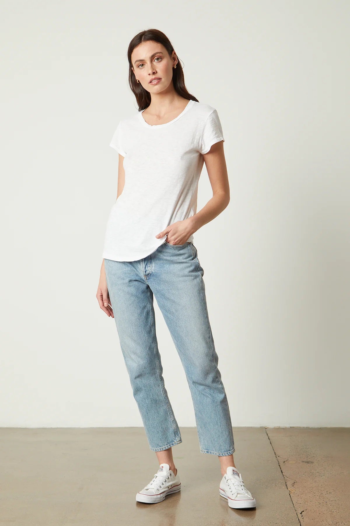 A woman with long brown hair stands sideways, looking towards the camera. She is wearing a hi-lo luxe essential beige VELVET - ODELIA CREW NECK COTTON SLUB TEE from VELVET BY GRAHAM &amp; SPENCER and dark blue jeans. Her left hand is in her pocket, and she stands against a plain white background.