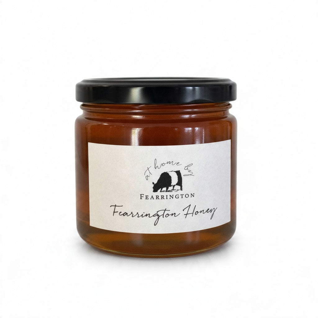 The Fearrington Signature Collection's hexagonal glass jar elegantly holds their Wildflower Honey, known for its raw quality. The label states: "Raw, Unfiltered. Handmade with care in our kitchens. May contain allergens. 282 grams, 10 oz." Inside is the finest golden honey from Fearrington House.