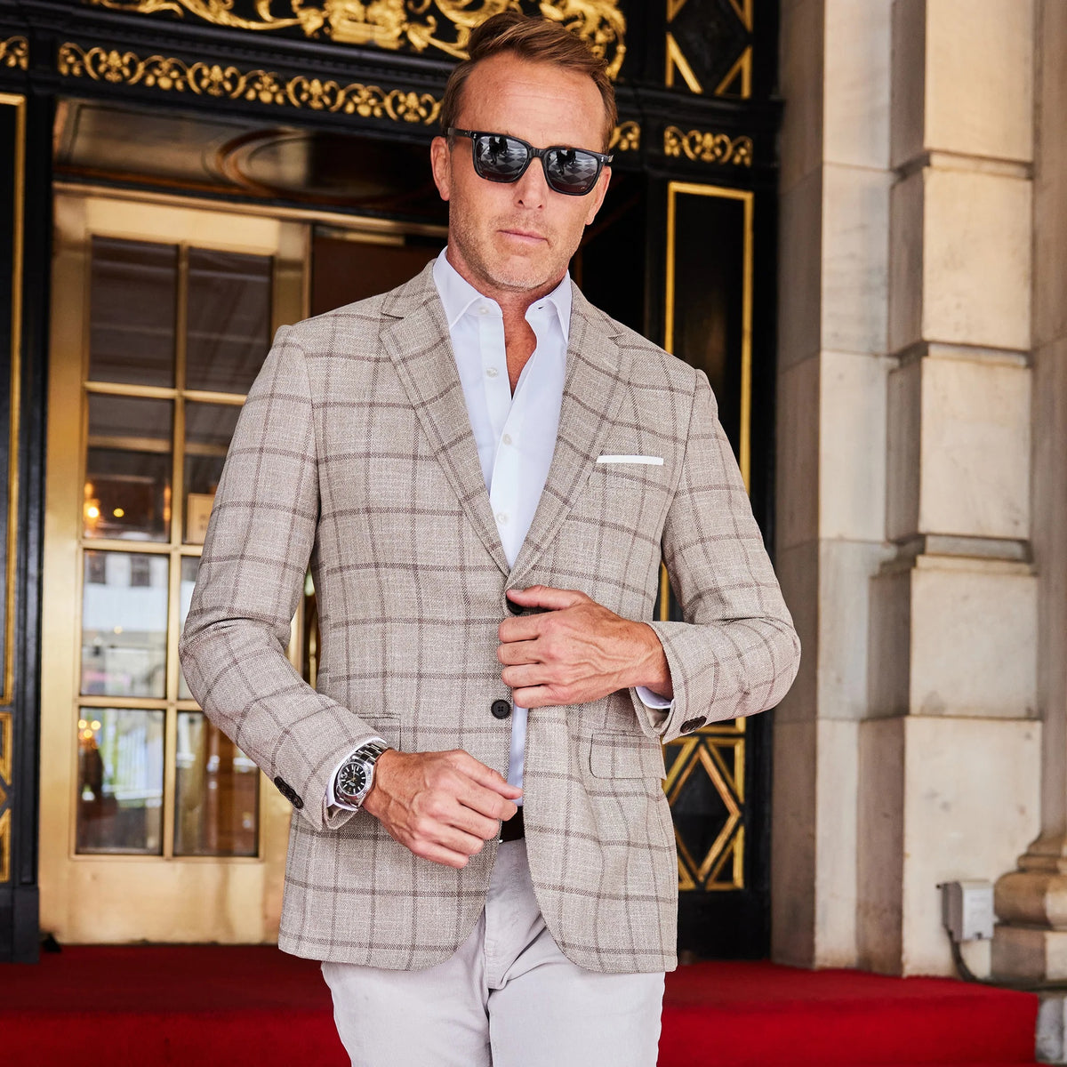 A man in the COLLARS AND CO MAVERICK TRAVEL BLAZER IN BEIGE WINDOWPANE and a white shirt stands in a doorway, holding sunglasses and the door, looking to the side with a serious expression.