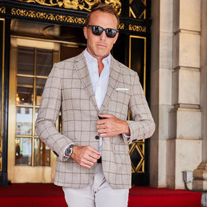 A man in the COLLARS AND CO MAVERICK TRAVEL BLAZER IN BEIGE WINDOWPANE and a white shirt stands in a doorway, holding sunglasses and the door, looking to the side with a serious expression.