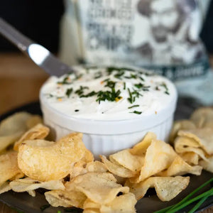 A bag of CAROLINA KETTLE - CREAM CHEESE AND CHIVE CHIPS by 1 IN 6 SNACKS features a vintage illustration of Sir Walter Raleigh. The enticing flavor combination is Cream Cheese & Chive. The packaging design is green with white and dark green text, and includes "Gluten Free" and "1 in 6 Snacks" labels.