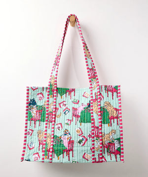 A woman in a white shirt and white pants smiles while holding the PRINTFRESH Mahjong Menagerie Traveler Tote—an eye-catching, quilted bag with colorful red, green, and blue designs—over her shoulder, standing against a plain light-colored background.