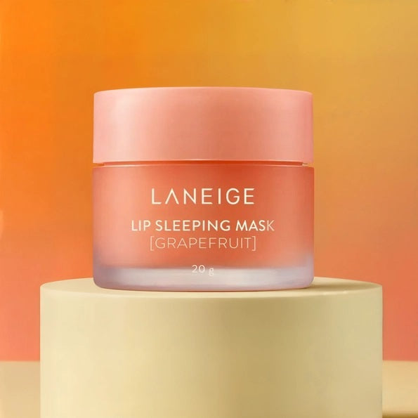 A jar of KOSMIC KBEAUTY SKINCARE’s LANEIGE - LIP SLEEPING MASK in Grapefruit rests on a beige pedestal against a warm orange and yellow gradient background, showcasing this nourishing overnight lip treatment.