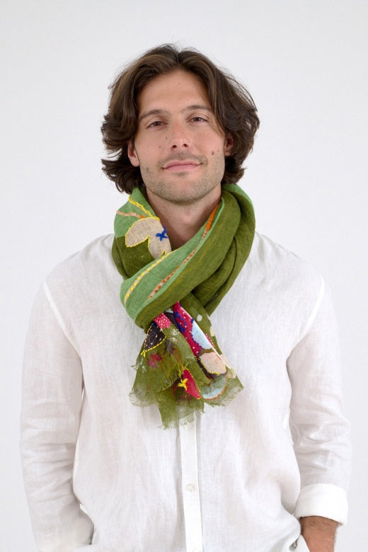 A man with wavy brown hair wears a white long-sleeve shirt, beige pants, and the STORIATIPIC - AZURE SCARF by STORIATIPIC, featuring colorful handmade embroidery, standing against a plain white background.