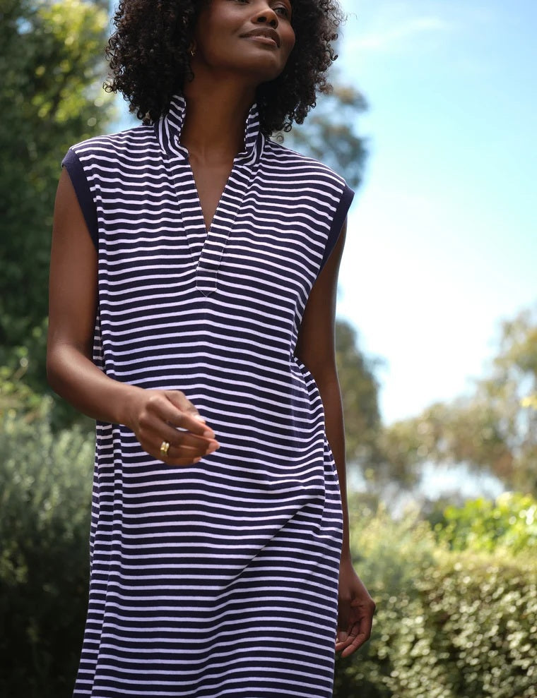 A person smiles outdoors, wearing the FRANK & EILEEN Mini Lily Popover Henley Tank Dress in Vintage Jersey Yacht Stripe, a sleeveless navy and white striped dress made in California. Lush greenery and a bright blue sky appear in the background.