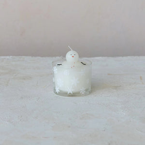 The CREATIVE COOP MELTING SNOWMAN CANDLE is a white holiday candle in a glass holder with snowflakes and a small snowman figure on top, set against a light, textured backdrop—perfect for festive winter decor.