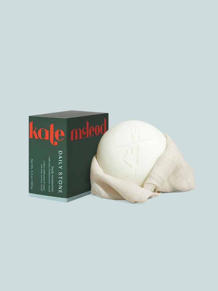 A cloth-wrapped soap bar sits next to a dark green box marked "KATE MCLEOD" in red and "Daily Stone" in white. The Body Stone features a raised logo design, reflecting the elegance of a luxury lotion bar against a light gray backdrop.