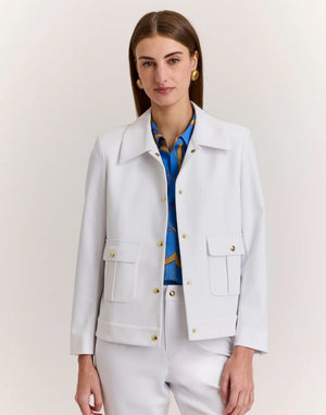 A woman with straight brown hair wears the HINSON WU - HANNAH TRAVELER'S TWILL JACKET with gold buttons over a blue patterned shirt and white pants, standing against a plain light background.