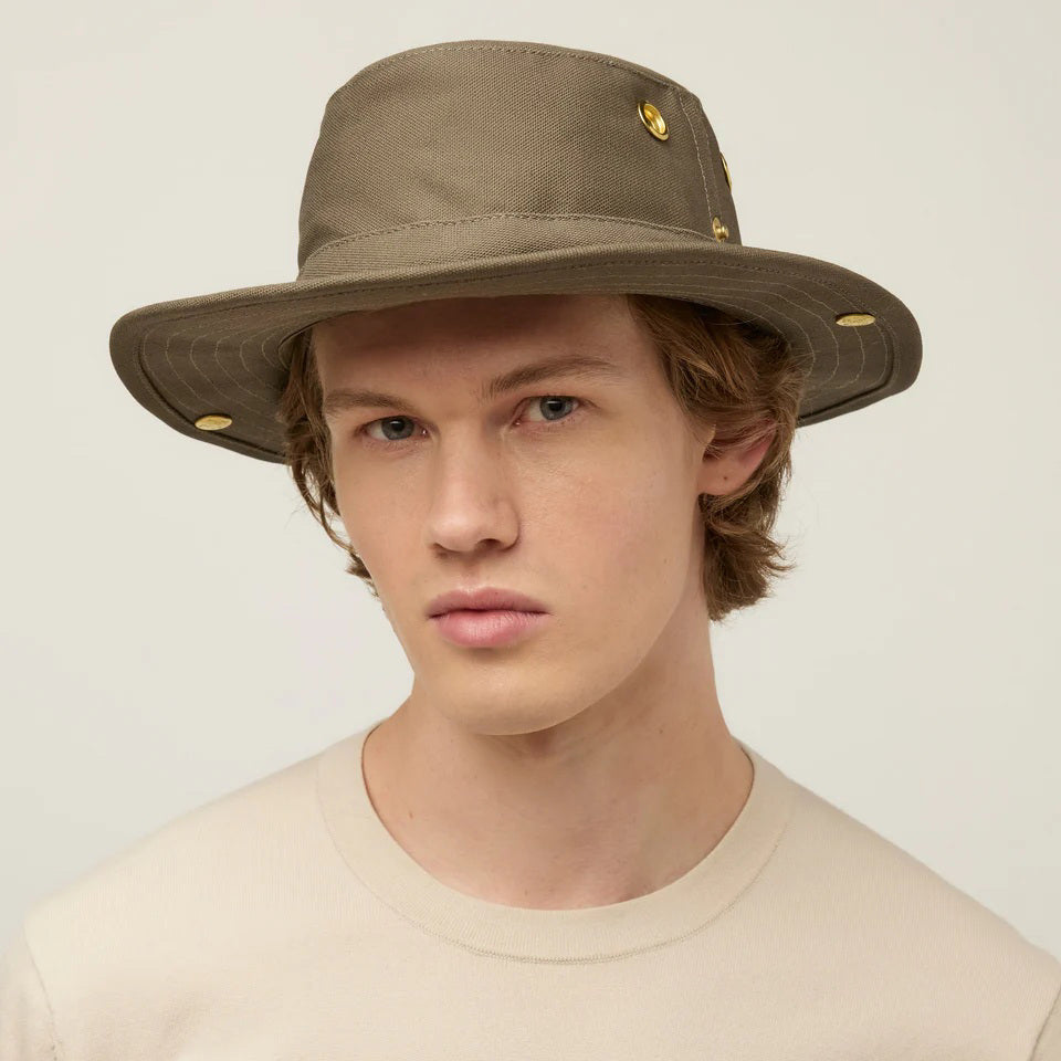 A young person with light skin and wavy brown hair wears a beige TILLEY - CLASSIC T3 HAT from TILLEY, featuring gold accents, paired with a beige shirt and looking at the camera against a plain light background.
