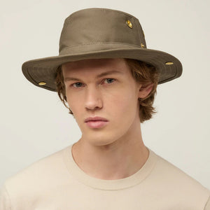 A young person with light skin and wavy brown hair wears a beige TILLEY - CLASSIC T3 HAT from TILLEY, featuring gold accents, paired with a beige shirt and looking at the camera against a plain light background.