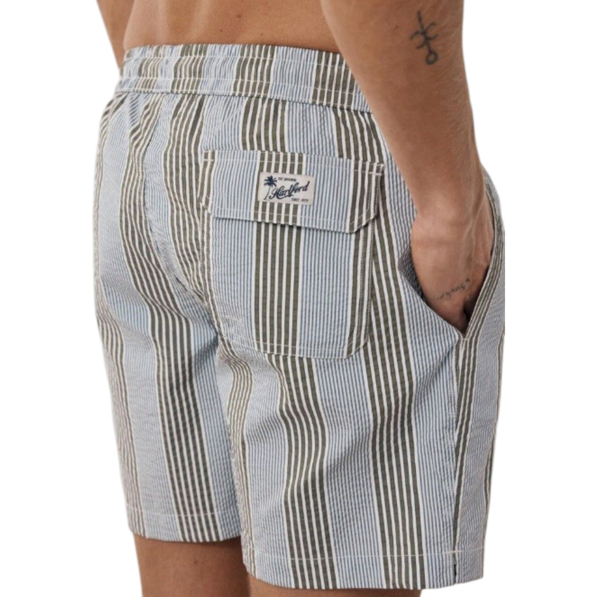 HARTFORD Seersucker Swim Trunks in green and blue stripes for men feature an elastic waistband, white drawstring, and olive accents. These mid-length swim shorts offer a classic look in textured seersucker fabric, shown on a light background.