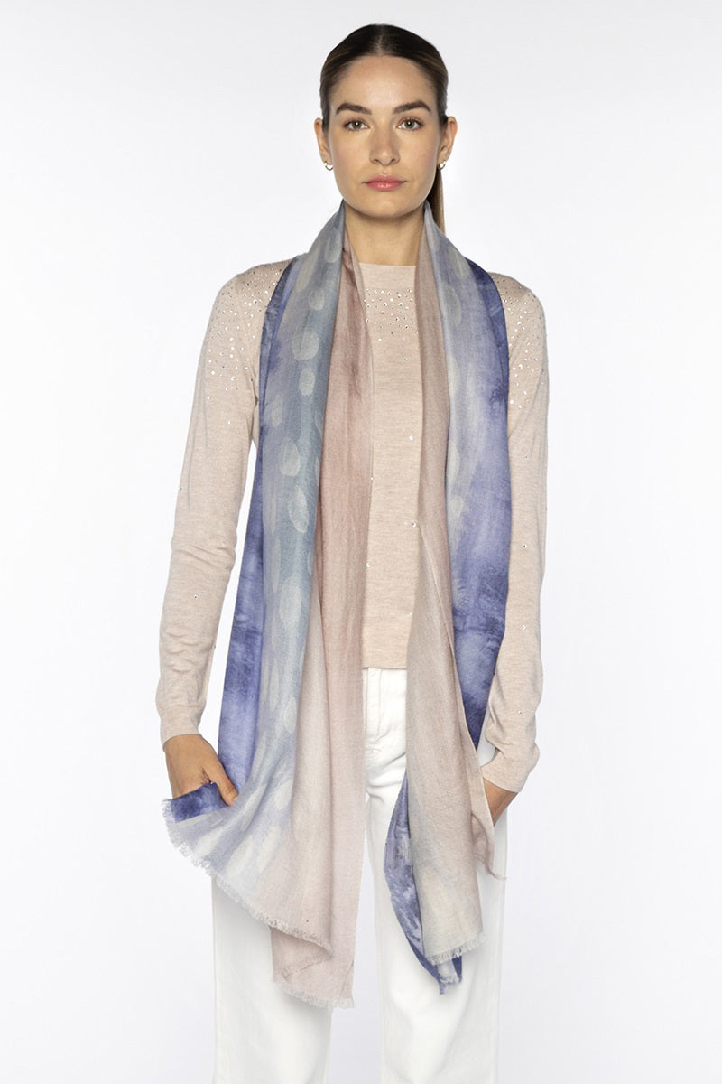A woman stands against a plain background wearing a light beige long-sleeve top, white pants, and a KINROSS CASHMERE Watercolor Dot Scarf in Navy Multi elegantly draped around her neck.