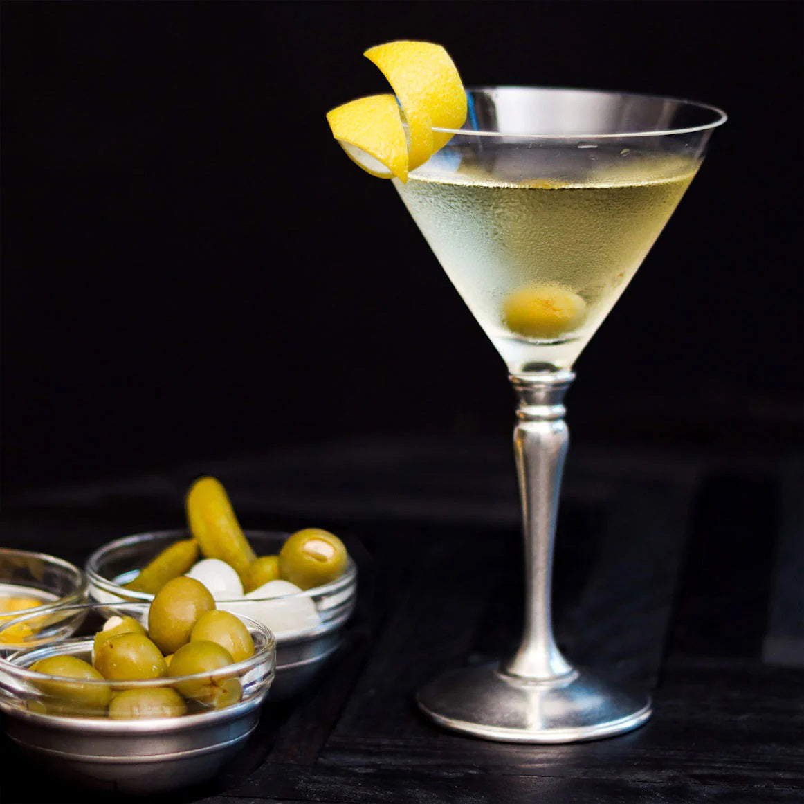 A martini with an olive and lemon twist in a MATCH - CLASSIC MARTINI GLASS by MATCH sits beside bowls of green olives, cocktail onions, and pickles against a dark background.