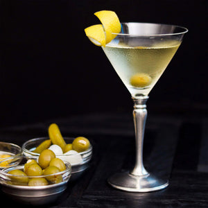 A martini with an olive and lemon twist in a MATCH - CLASSIC MARTINI GLASS by MATCH sits beside bowls of green olives, cocktail onions, and pickles against a dark background.