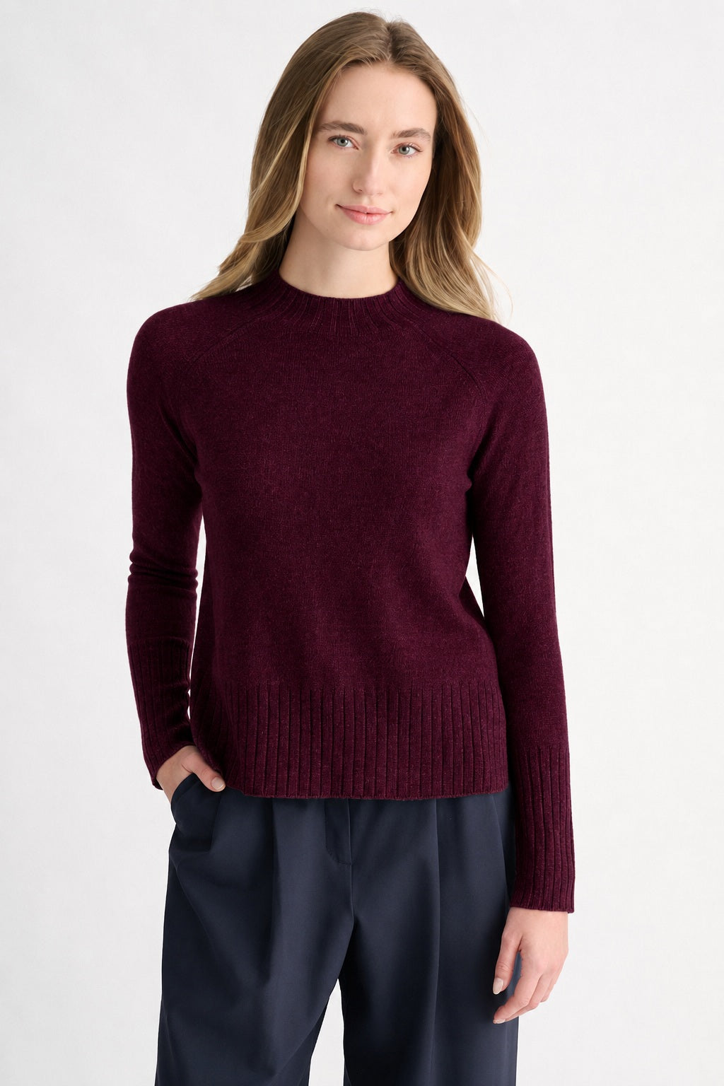 A person with long, light brown hair wears the KINROSS CASHMERE Hi Low Rib Trim Funnel Cashmere Sweater in Cordovan and dark pants, standing against a plain light background with one hand in their pocket, looking at the camera and smiling slightly.