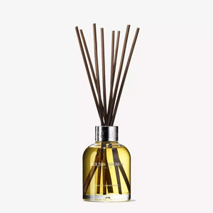A clear glass bottle filled with yellow liquid, labeled "MOLTON BROWN - ORANGE AND BERGAMOT DIFFUSER," features several long, thin, dark brown diffuser reeds extending from its neck. The reed diffuser boasts a hint of Sevillian orange and neroli. A metallic ring at its opening gives it an elegant and classy appearance.