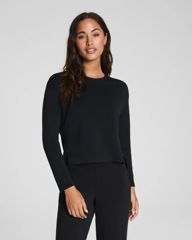 A woman with long dark hair wears the SPANX AirEssentials Relaxed Long Sleeve Top and matching black pants, standing against a plain light gray background and looking forward with a neutral expression.