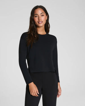 A woman with long dark hair wears the SPANX AirEssentials Relaxed Long Sleeve Top and matching black pants, standing against a plain light gray background and looking forward with a neutral expression.