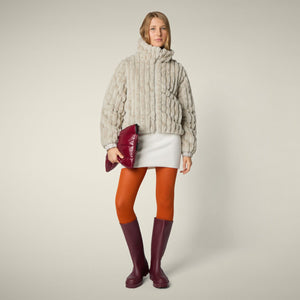 A woman models the SAVE THE DUCK - ELINA REVERSIBLE JACKET in beige fur, styled with a white skirt, orange tights, burgundy boots, and a matching clutch, standing against a plain light background.
