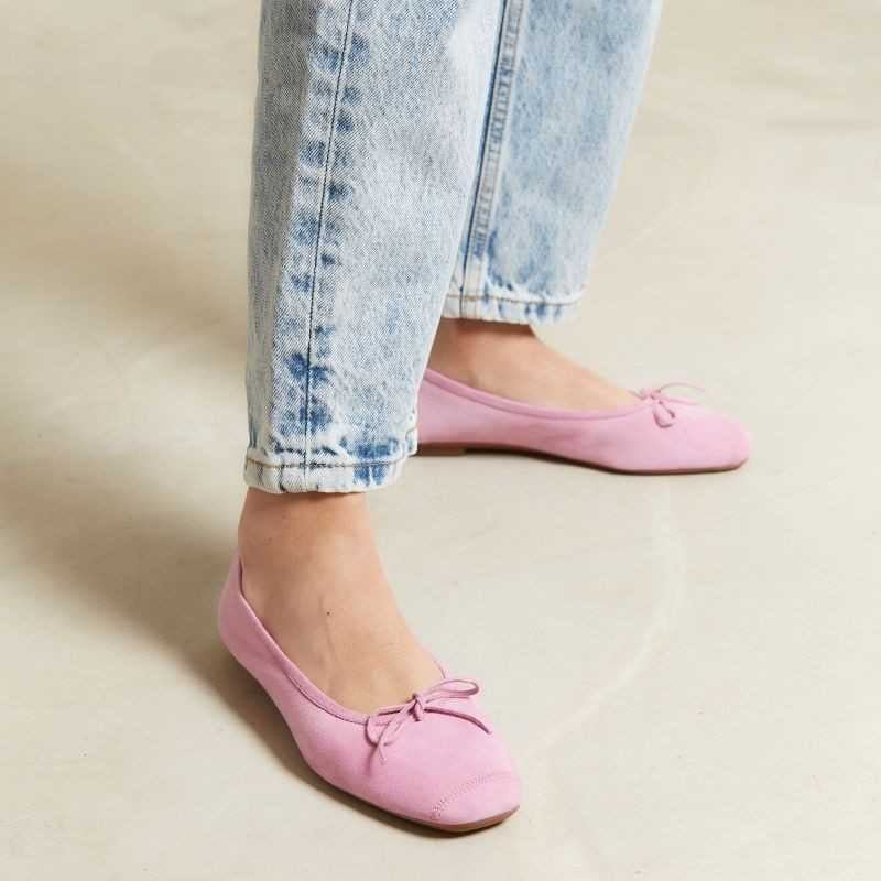 A close-up of the REQINS - HARMONY SUEDE BALLERINA FLATS IN PINK, featuring a bow detail on the front, photographed on a light surface—an elegant update to this classic women’s shoe by REQINS.