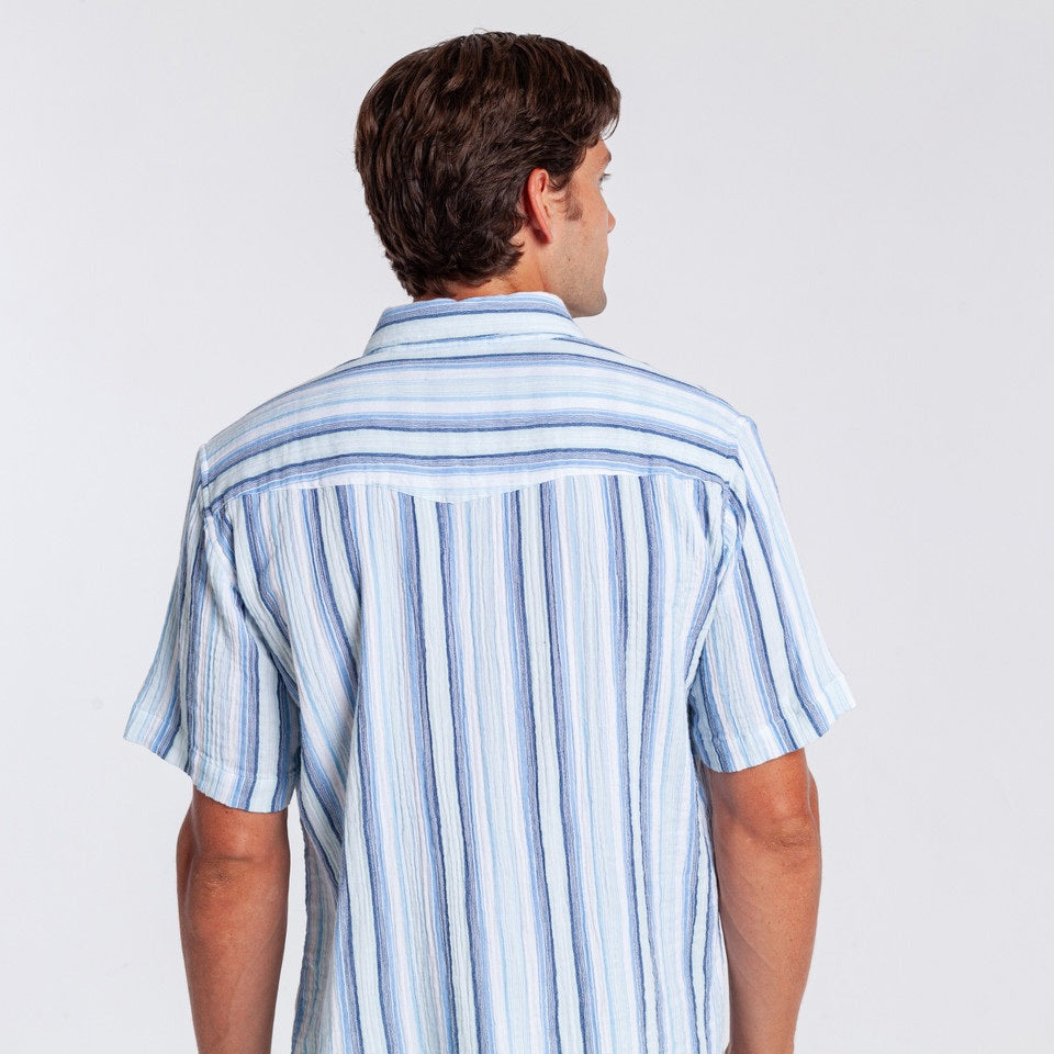 A man wearing the CRIQUET - THE BEACHCOMBER SHIRT IN BLUE by CRIQUET, paired with light beige shorts, stands against a plain white background, looking to his left with one hand in his pocket.