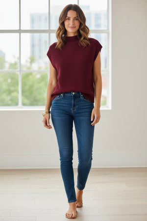 A woman is seen standing against a plain white background, smiling gently with one hand relaxed by her side, wearing the KINROSS CASHMERE Cap Sleeve Cashmere Rib Funnel in Cordovan from KINROSS, paired with blue jeans.