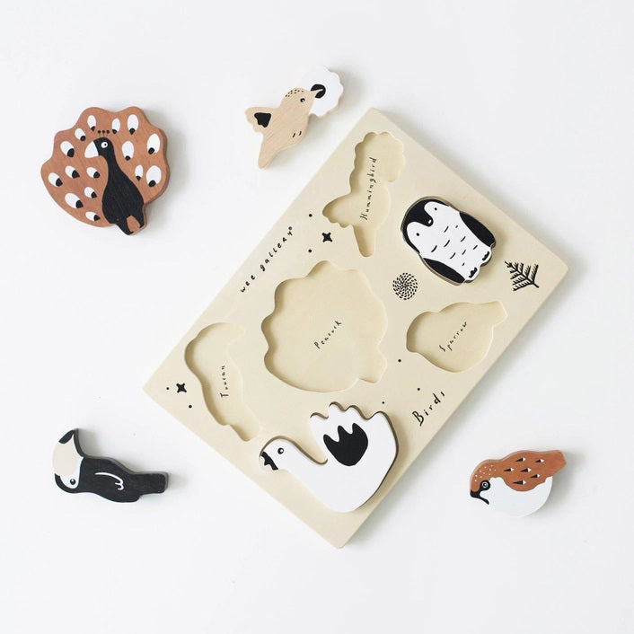 The WEE GALLERY - BIRDS TRAY PUZZLE is a wooden board featuring six bird-shaped pieces, including a toucan, peacock, penguin, swan, sparrow, and a small bird on a branch. This educational puzzle offers fun with its simple colors and shapes. The board is labeled with "wee gallery" and "Birds".