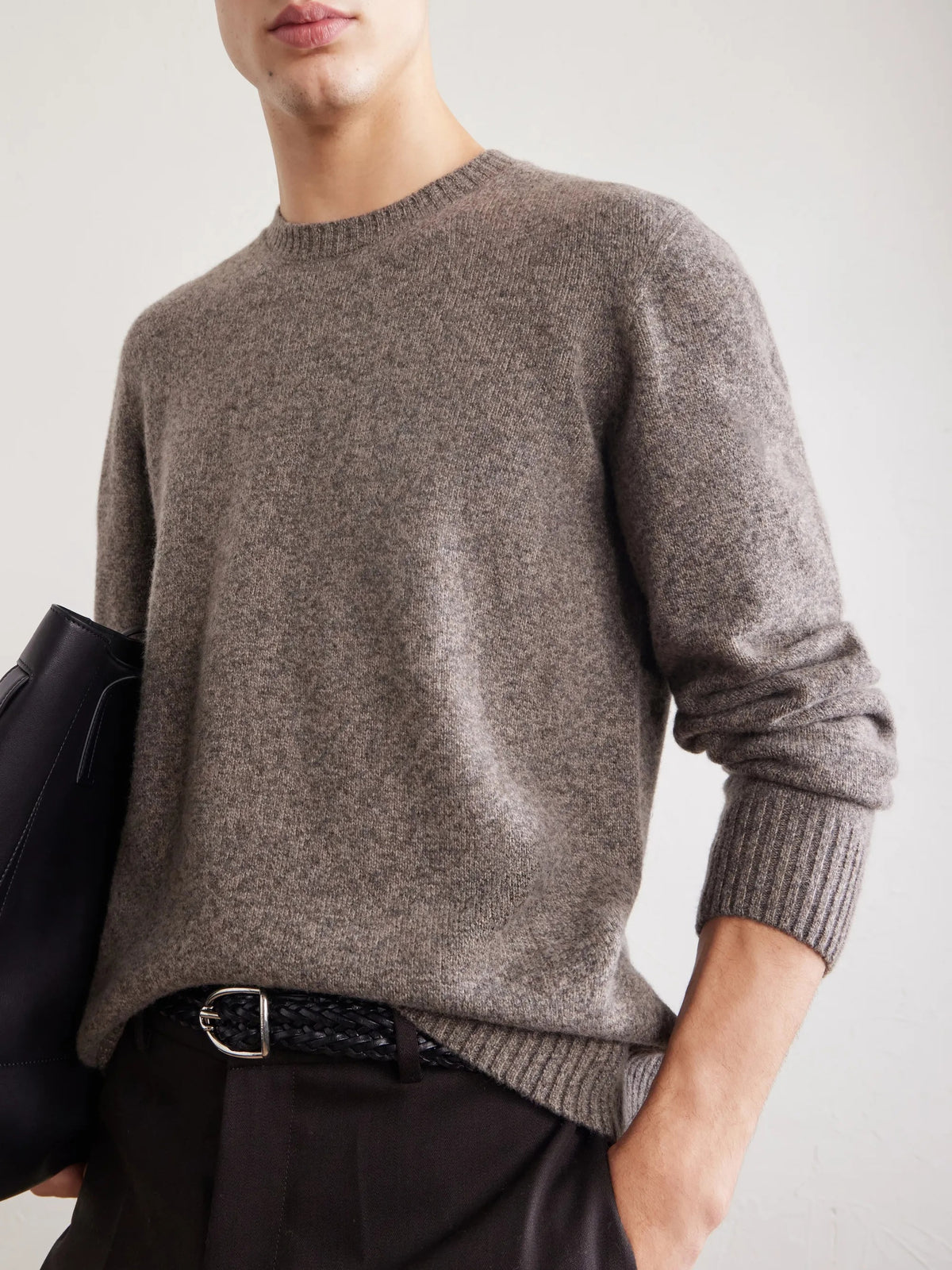 A person wearing the HARTFORD Seamless Wool Crew Pullover in Mushroom, an Italian-made sweater, paired with dark pants, is seated with one arm resting on their knee against a plain light background.
