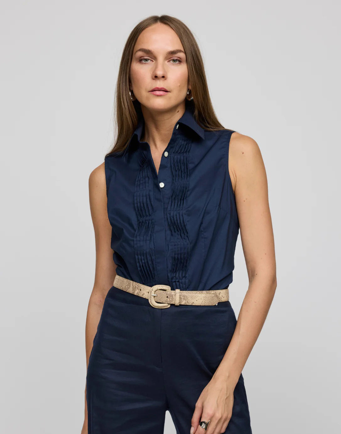 A woman with long hair wears the HINSON WU Lilith Sleeveless Shirt, made from stretch polished cotton with pleats and a beige belt, against a gray background. She exudes confidence as she gazes at the camera, highlighting the shirt&#39;s versatility.