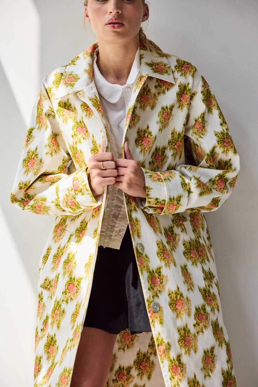 A person stands on grass before green bushes, wearing HOPE FOR FLOWERS' pink Warped Ikat coat over a white shirt and matching shorts with black sandals, looking to the side and highlighting the brand's commitment to ethical fashion.
