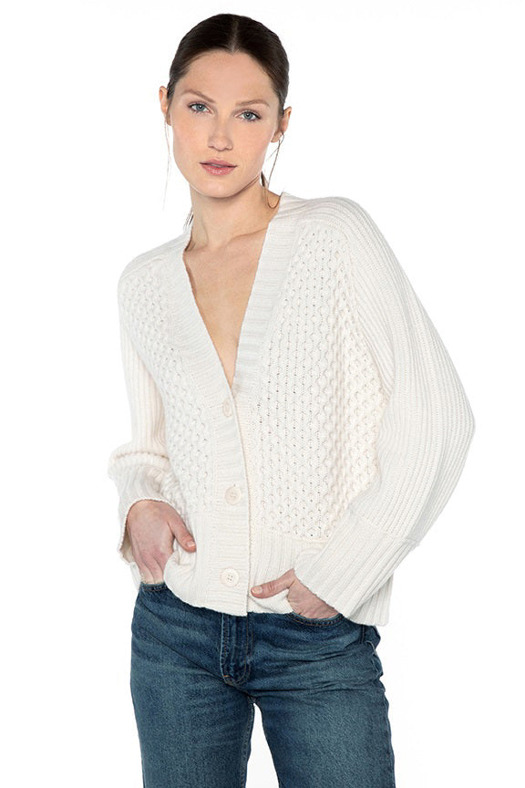 A person with long hair pulled back is wearing a KINROSS CASHMERE LUXE MIXED STITCH CARDIGAN from KINROSS, made of 100% cashmere, featuring a textured front and ribbed sleeves. The white cardigan is paired with blue jeans and is buttoned except for the top and bottom buttons. They have their hands in their pockets and are posing against a white background.
