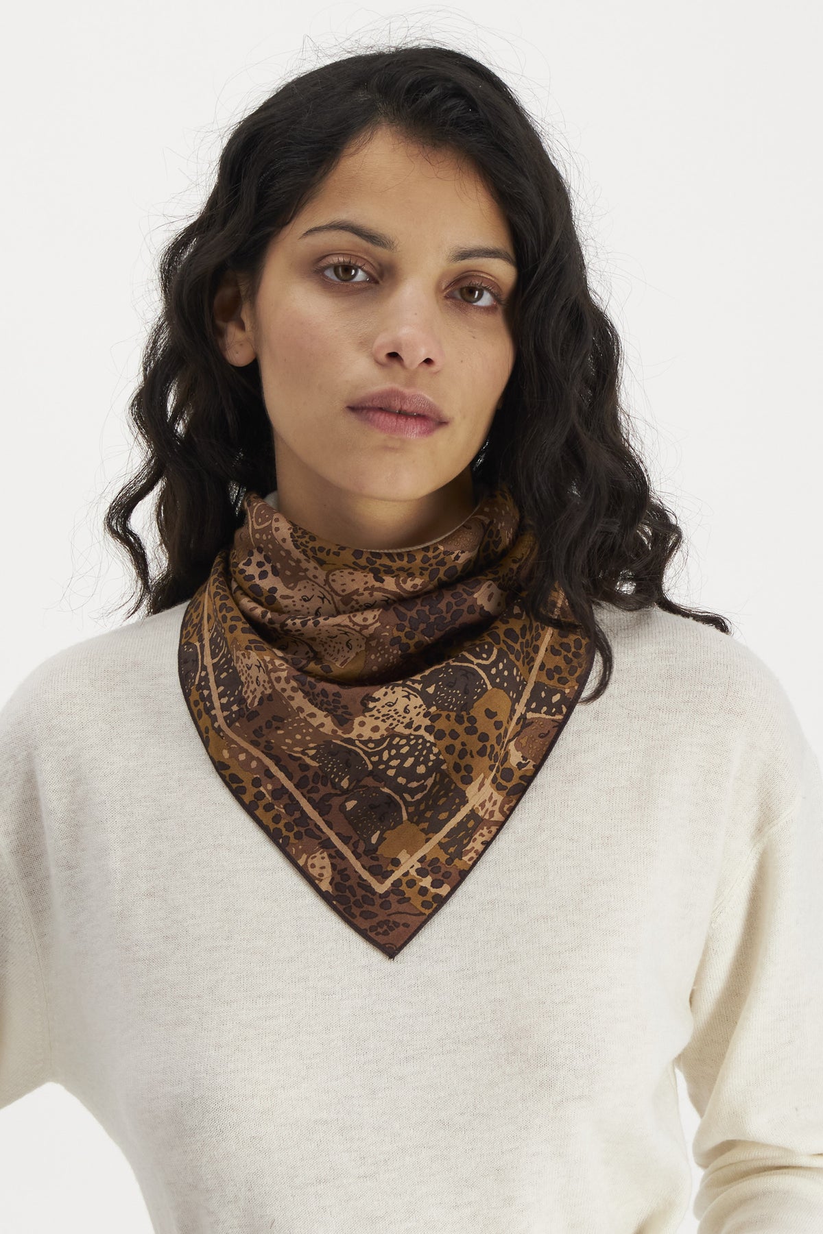 A person with long, wavy dark hair wears a light cream sweater and an intricately patterned brown fashion accessory from INOUI around their neck. The INOUI EDITIONS TETRIS SQUARE COTTON/SILK SCARF, made from soft cotton silk, features various shades of brown with detailed floral and geometric motifs. They stand against a plain white background.