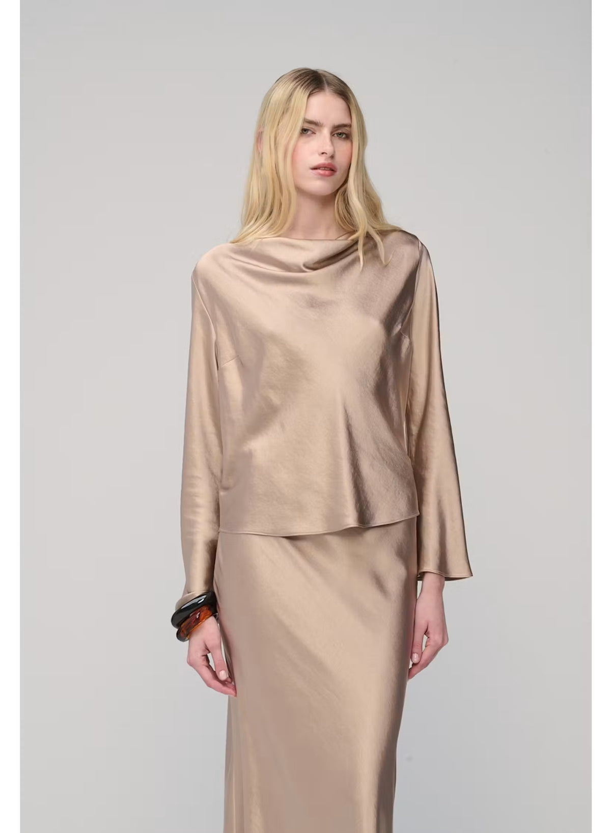 A woman with long blonde hair poses against a plain light gray background, wearing the MILVA SATIN BLOUSE IN SAND by MILVA—a shiny, champagne-beige long-sleeve blouse featuring a chic cowl neckline—paired with a matching skirt and accessorized with bracelets on her left wrist.
