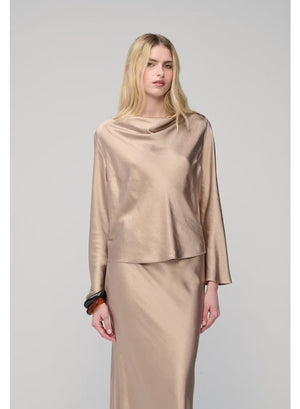 A woman with long blonde hair poses against a plain light gray background, wearing the MILVA SATIN BLOUSE IN SAND by MILVA—a shiny, champagne-beige long-sleeve blouse featuring a chic cowl neckline—paired with a matching skirt and accessorized with bracelets on her left wrist.
