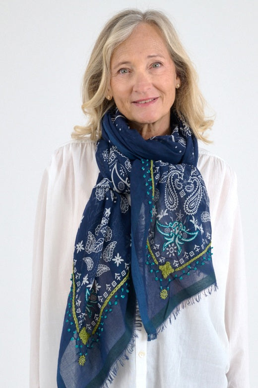A person with long gray hair displays the STORIATIPIC - KIKI SCARF IN MARINE by STORIATIPIC, a large sheer blue linen scarf with white floral and paisley patterns and delicate handmade embroidery, against a plain white background.