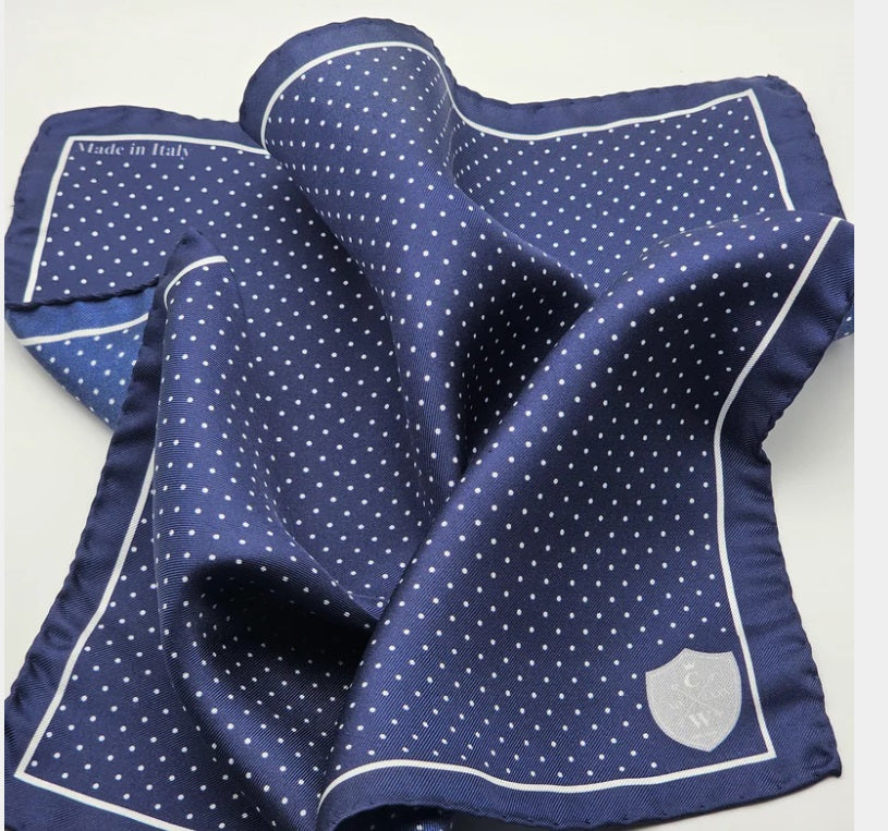The CLIFTON WILSON - NAVY &amp; WHITE POLKA DOT SILK POCKET SQUARE features a navy silk base with a white polka dot grid and thin white border, "Made in Italy" in the top left, and a small white shield emblem in the bottom right. Brand: CLIFTON WILSON.
