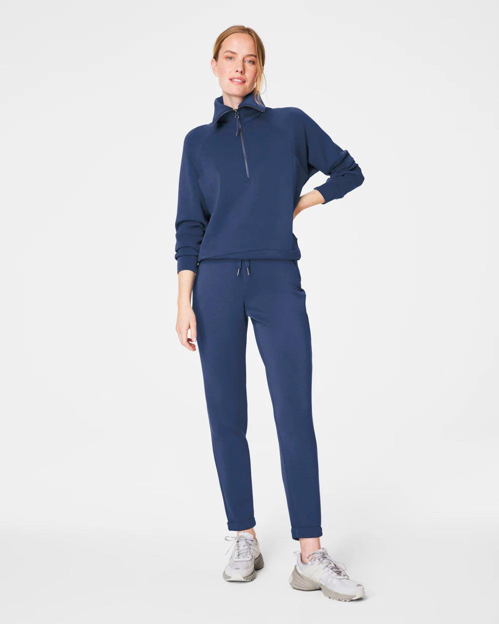 A person stands against a white background, smiling and looking down with one hand touching their hair. They are wearing a matching light beige SPANX AIRESSENTIALS TAPERED PANT and sweatshirt set made from ultra drapey, lightweight spacer fabric, paired with white sneakers.